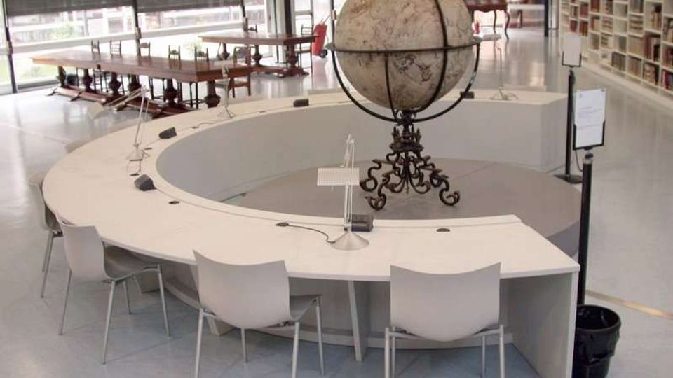 Sala di biblioteca o studio moderna con grande tavolo circolare bianco e sedie, al centro un globo terrestre antico su supporto decorato. Sullo sfondo ci sono lunghe finestre con tende, tavoli in legno e scaffali pieni di libri.