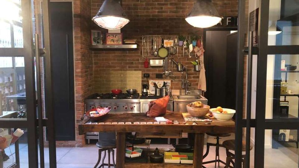 Cucina in stile industriale con pareti in mattoni a vista, grande tavolo in legno al centro e due lampade metalliche a sospensione. Sul fondo si vedono fornello e lavello con mensole e griglia portaoggetti, incorniciati da una struttura in vetro e metallo.