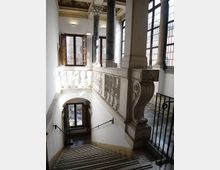 Interno di un edificio storico con scala in pietra che scende verso un pianerottolo, affiancata da una balaustra in marmo scolpito. Grandi finestre con griglie e colonne in marmo illuminano l’ambiente, con soffitto decorato e pavimento a mosaico visibili al piano superiore.