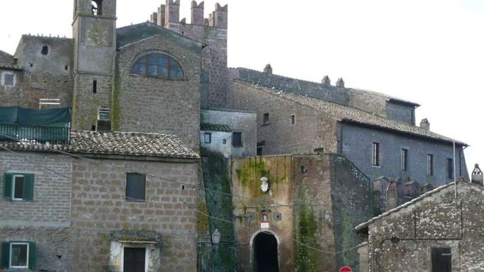 Complesso storico in pietra e mattoni con torre campanaria, edifici addossati e tetti in coppi, visto da una piazza. Al centro si apre un grande arco d’ingresso su un passaggio in salita, con mura esterne segnate da muschio.