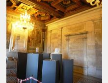 Interno di una sala storica con pareti in pietra incise, soffitto a cassettoni in legno dorato e grandi lampadari. Al centro sono presenti piedistalli scuri con frammenti scultorei e pavimento a motivi geometrici.