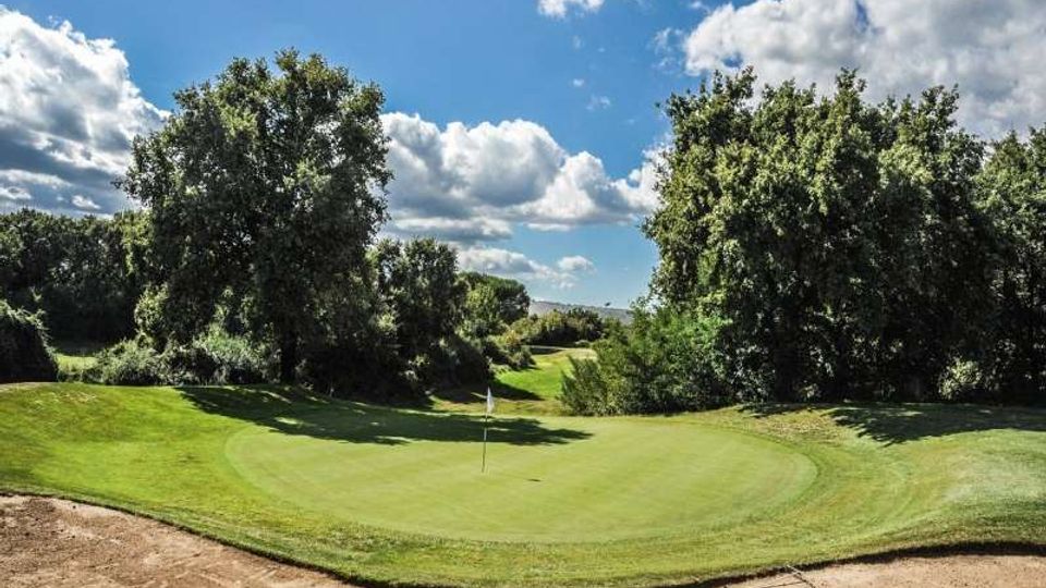 Campo da golf con green circolare e bandierina al centro, circondato da alberi e vegetazione. In primo piano si vede un bunker di sabbia, con cielo azzurro e nuvole sullo sfondo.