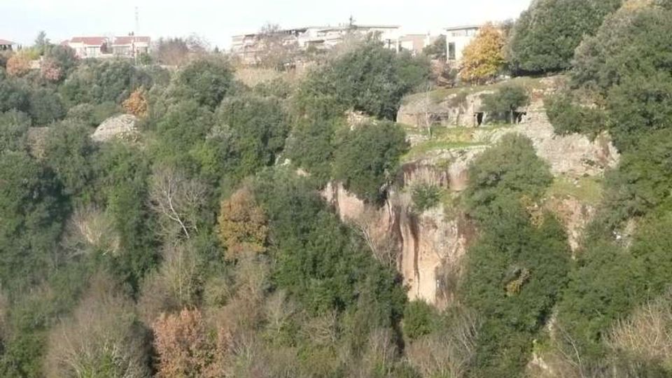 Veduta di una gola boschiva con pareti di roccia e vegetazione fitta, con alberi dai colori autunnali. In alto si vedono alcune case e costruzioni sul bordo della scarpata.