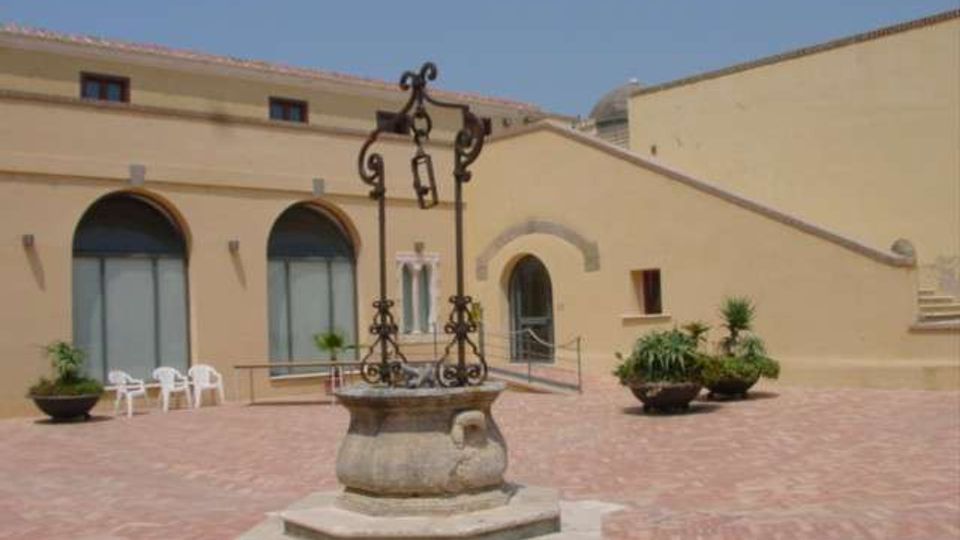 Cortile lastricato in mattoni con un pozzo in pietra al centro e struttura in ferro battuto. Sullo sfondo edifici color sabbia con grandi archi vetrati, una porta ad arco e alcune piante in vaso lungo i lati.