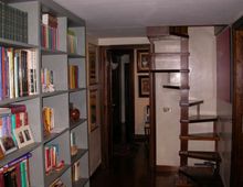 Interno di un’abitazione con corridoio stretto: a sinistra una grande libreria a scaffali pieni di libri e oggetti, a destra una scala in legno con gradini aperti. Pareti con cornici e porte in legno, pavimento scuro e soffitto basso.
