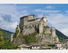 Castello medievale in pietra arroccato su uno sperone roccioso, con mura fortificate, torrioni e una torre con orologio. In primo piano si vedono i tetti di un borgo, con colline verdi e cielo parzialmente nuvoloso sullo sfondo.