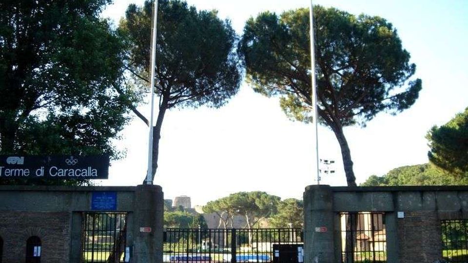 Ingresso recintato delle Terme di Caracalla con cancelli in metallo e pilastri in muratura, affiancato da due alti pini marittimi e pennoni. Sullo sfondo si vedono aree verdi e le rovine archeologiche all’interno del sito.