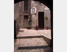 Cortile acciottolato visto da sotto un arco, con facciata di edificio in pietra e intonaco, porta in legno scuro e finestra con persiane. Sul muro c’è un’edicola con immagine e un cartello “Via Castello”, con vasi di fiori e piante disposti vicino all’ingresso.