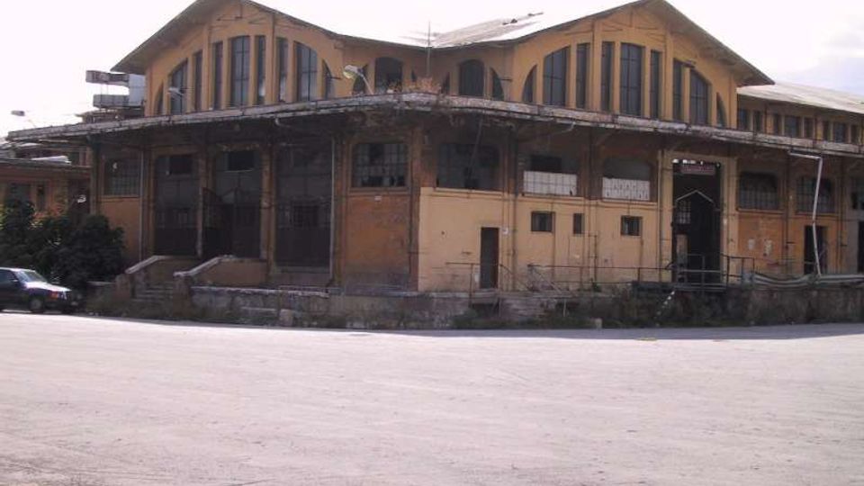 Grande edificio industriale in mattoni con tetto a capanna e ampie finestre ad arco, affacciato su un ampio piazzale asfaltato. Una tettoia sporgente corre lungo la facciata con portoni e rampe di carico.