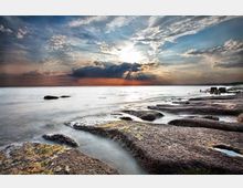 Costa rocciosa sul mare con ampie piattaforme di scogli scuri e pozze d’acqua tra le rocce, che si estendono verso l’orizzonte. Sullo sfondo il sole basso filtra tra nuvole dense, illuminando la superficie marina e una linea di costa lontana.