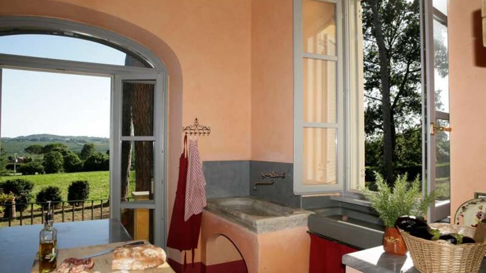 Interno di una cucina rustica con pareti color pesca, lavello in pietra e grandi finestre ad arco con infissi chiari aperte su un paesaggio di campagna verde. Piano di lavoro con alimenti e bottiglia, e vista esterna con alberi e colline.