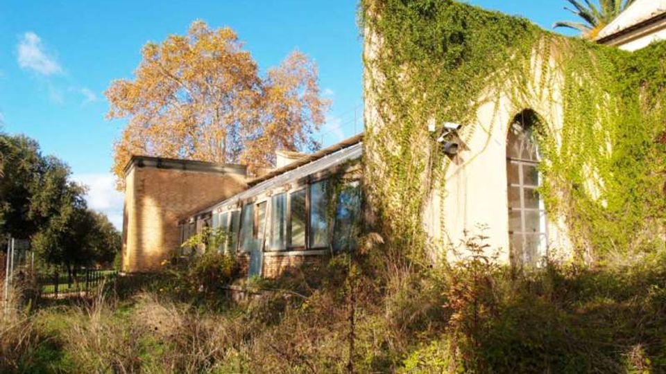 Edificio rurale in parte abbandonato, con pareti esterne ricoperte di edera e una grande finestra ad arco; sul lato sinistro si vede un’ala con ampie vetrate. In primo piano c’è vegetazione alta e incolta, con alberi sullo sfondo sotto un cielo sereno.