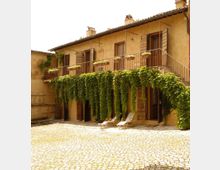 Cortile lastricato in pietra davanti a un edificio rustico a due piani con intonaco beige, balcone con ringhiera metallica e persiane in legno. Tralci di vite pendono dal balcone fino al piano terra e sono presenti sedie a sdraio vicino alle porte.