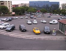 Ampia rotatoria adibita a parcheggio, con numerose auto disposte lungo la curva e carreggiata asfaltata in primo piano. Sullo sfondo si vedono alberi, edifici residenziali e un grande edificio moderno con facciata in vetro, oltre a un’area con auto parcheggiate lungo la strada.