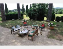 Area salotto all’aperto su ghiaia in un giardino, con sedute in legno e tavolini attorno a un tavolo con tovaglia. Intorno ci sono grandi pini, siepi e vasi di ortensie, con vista su un prato e colline sullo sfondo.