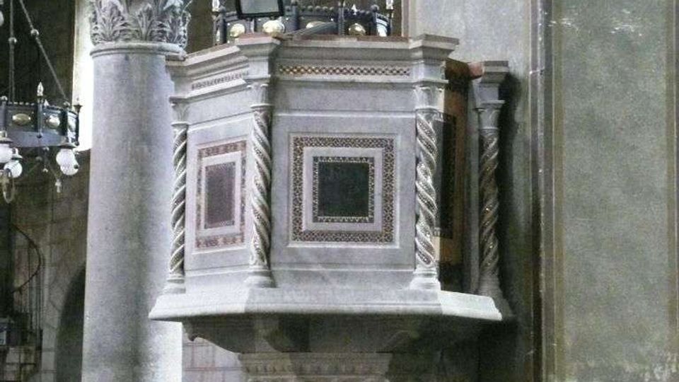 Interno di una chiesa storica con pulpito in marmo decorato da pannelli geometrici e colonne tortili, addossato a un pilastro. Si vedono archi affrescati, colonne in pietra e lampadari sospesi lungo la navata.