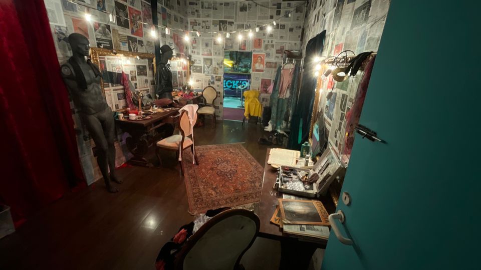 Interno di un camerino teatrale con pareti e soffitto rivestiti di pagine di giornale e una fila di lampadine lungo i lati. Specchi con cornici dorate, tavoli da trucco pieni di oggetti, manichini, sedie e un tappeto centrale conducono a una porta in fondo che si apre su un corridoio illuminato.