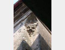 Dettaglio dal basso dell’angolo di un edificio in pietra, con una piccola scultura in rilievo raffigurante una testa grottesca incassata nella muratura. In alto si vede la gronda con tegole e una porzione di cielo.
