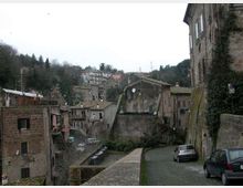 Vista di un borgo collinare con case in pietra e tetti in coppi, disposto su più livelli lungo il pendio. In primo piano si vede una strada stretta con un muretto in pietra e alcune auto parcheggiate, mentre sullo sfondo ci sono alberi e abitazioni sparse sulla collina.