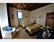Camera da letto con due letti singoli affiancati, soffitto con travi in legno a vista e pavimento in piastrelle. Una finestra con tenda chiara illumina la stanza, arredata con poltrona bianca, comodino con lampada, quadri alle pareti e porta in legno.
