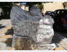 Scultura in pietra chiara a forma di uccello stilizzato con ali spiegate su basamento, collocata in una piccola area pavimentata all’aperto. Sullo sfondo si vedono un muro e edifici, alberi che fanno ombra e alcune auto parcheggiate.