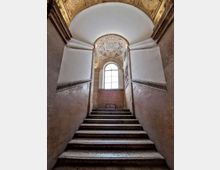 Scala interna in pietra che sale verso una grande finestra ad arco, con pareti intonacate e fascia decorativa orizzontale. In alto, soffitto a volta con stucchi e un medaglione circolare ornato.
