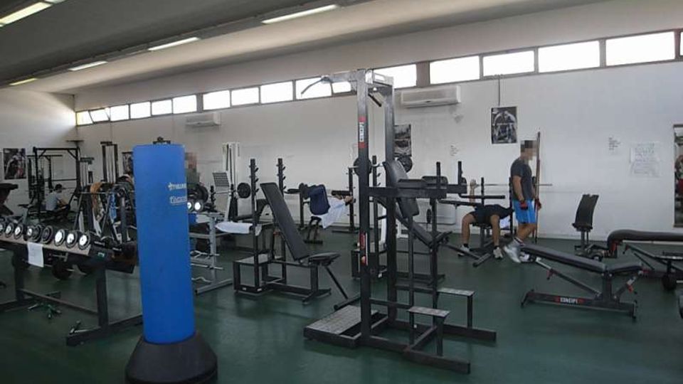Interno di una palestra con soffitto alto e finestre a nastro, pavimento verde e pareti bianche. Sono visibili panche, rastrelliere per pesi, macchine da allenamento e un sacco da boxe blu al centro della sala.