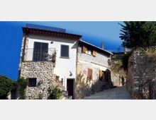 Edifici in pietra e intonaco bianco su una strada in salita, con persiane scure, balcone con ringhiera in ferro e finestre con scuri in legno. Il contesto è un borgo collinare con muri in pietra e vegetazione ai lati sotto un cielo limpido.