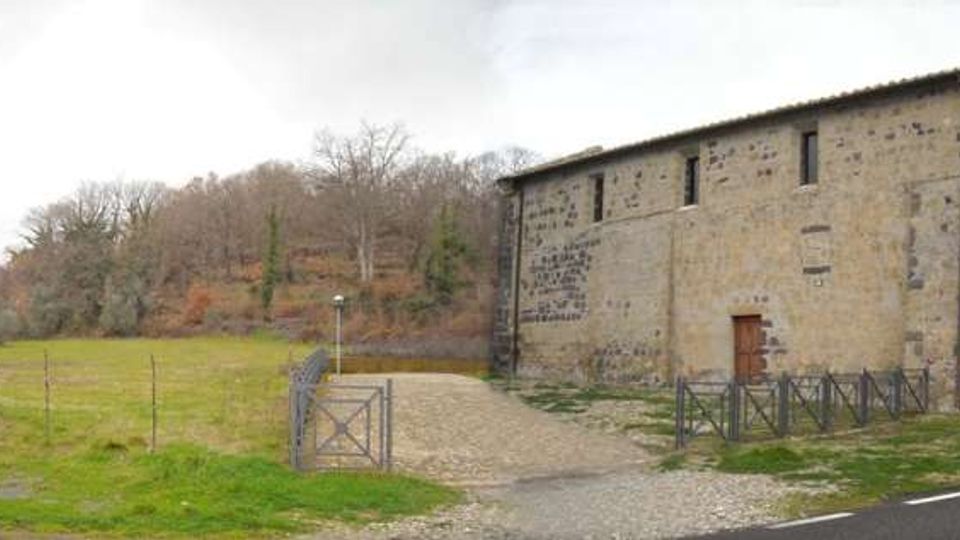 Edificio rurale in pietra con tetto a falde e piccole finestre, affacciato su un prato aperto. Un vialetto di ghiaia con cancello metallico conduce all’ingresso, con recinzione e alberi su una collina sullo sfondo.