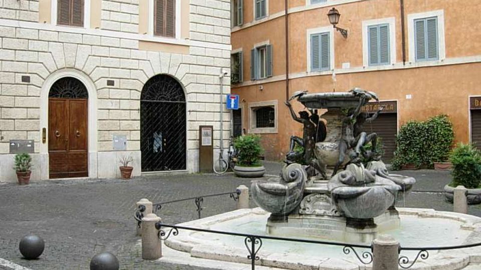 Piccola piazza lastricata con una fontana in pietra al centro, decorata con figure scolpite e vasche circolari con acqua. Intorno si vedono palazzi storici intonacati con finestre e portoni ad arco, lampioni a parete e dissuasori sferici lungo il bordo della piazza.