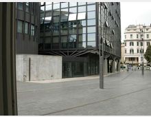 Ingresso di un moderno edificio per uffici con ampia facciata in vetro e struttura metallica, affacciato su una piazza pavimentata in cemento con pali di illuminazione. Sullo sfondo si vedono palazzi residenziali più tradizionali lungo la strada.