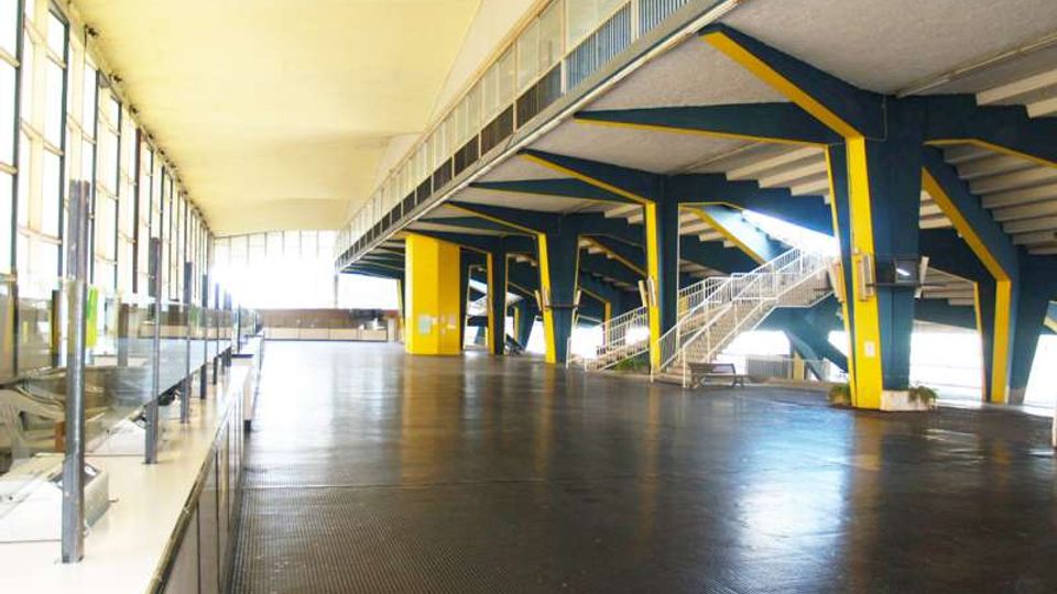 Ampio atrio interno di una struttura sportiva, con grande pavimentazione scura e colonne e travi in acciaio verniciate blu e giallo. Sulla sinistra si estende una parete di vetrate continue, mentre a destra si vedono gradinate e scale metalliche che conducono ai livelli superiori.