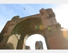 Veduta dal basso di un grande arco in muratura di un antico rudere, con pietre consumate dal tempo e un’ampia apertura centrale. Sullo sfondo si vede il cielo luminoso con riflessi di luce e alcune parti del manufatto mancanti o spezzate.