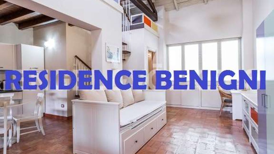 Soggiorno luminoso in open space con pavimento in cotto e soffitto alto con travi a vista, arredato con divano bianco e zona pranzo con tavolo e sedie. Sul fondo ampie porte-finestre vetrate e mobili bianchi lungo la parete.