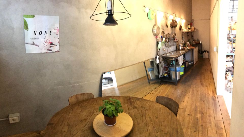 Interno di un laboratorio cucina dal design industriale, con pavimento in legno e pareti grezze. In primo piano c’è un tavolo rotondo in legno con una piccola pianta, mentre sul fondo si vede una lunga postazione con lavelli e utensili appesi, illuminata da lampade a sospensione.