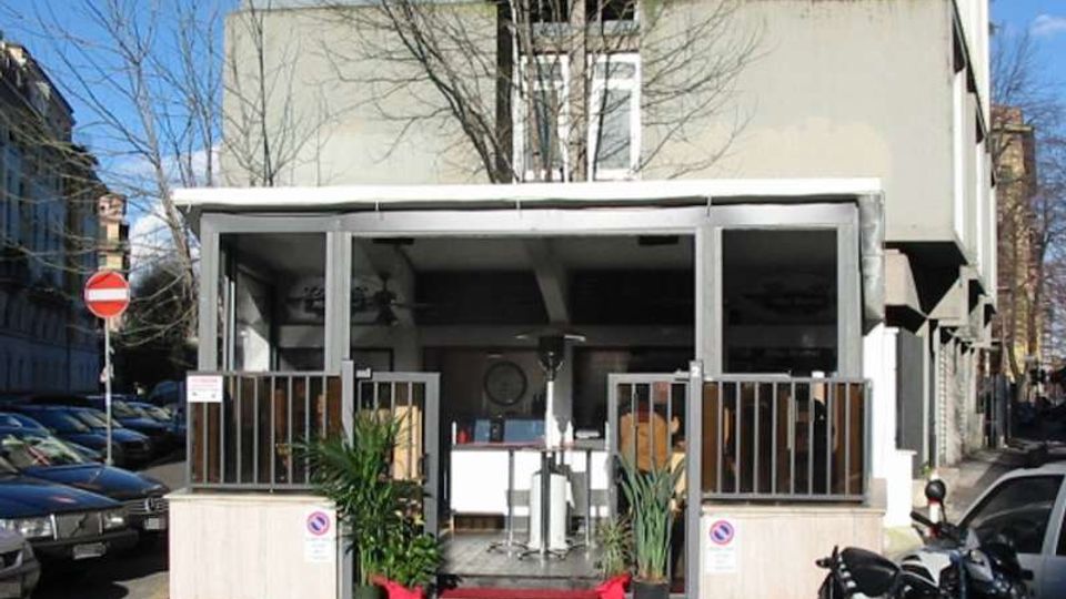 Ingresso di un edificio moderno su strada, con una veranda coperta e recintata da ringhiere metalliche e alcune piante in vaso ai lati. Sullo sfondo si vede la facciata chiara dell’edificio e, ai lati, la strada con auto parcheggiate e un segnale di divieto di accesso.