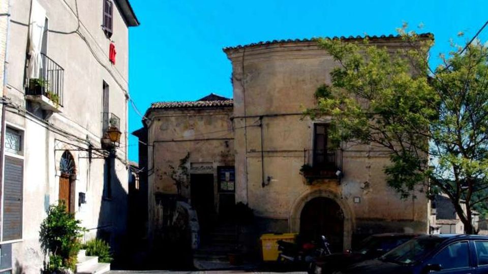 Piazzetta urbana con edifici in muratura intonacata: a sinistra una casa con balconi e persiane, al centro un edificio storico con portale ad arco e piccola loggia. In primo piano si vedono auto parcheggiate e un albero sul lato destro, sotto un cielo sereno.
