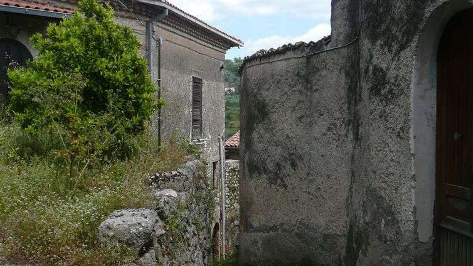 Vicolo stretto in un borgo con muri in pietra e intonaco, gradini in cemento e una lanterna in ferro battuto fissata alla parete. Ai lati si vedono case con tetti in tegole rosse, vegetazione spontanea e cielo azzurro sullo sfondo.