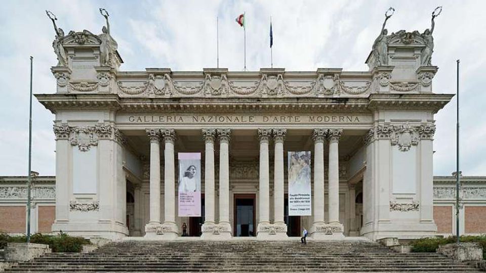 Facciata monumentale di un museo con ampio portico neoclassico sostenuto da colonne e scalinata frontale; in alto sono visibili decorazioni scultoree e bandiere. Sopra l’ingresso compare l’insegna “Galleria Nazionale d’Arte Moderna”, con grandi banner espositivi ai lati del portale.