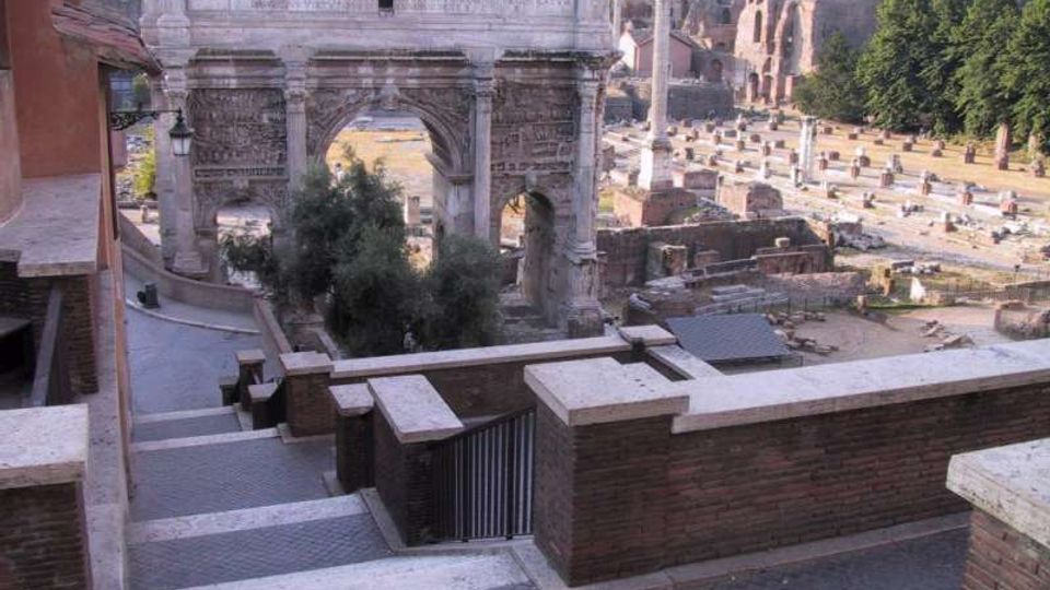 Veduta dall’alto di un arco monumentale romano in pietra con tre fornici e iscrizioni, affacciato su un’area archeologica con resti di edifici e colonne. In primo piano si vede una terrazza con muretti in mattoni e una scalinata pavimentata.