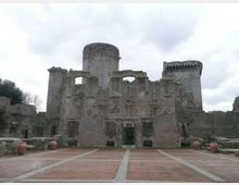Rovine di un castello in pietra con muri parzialmente crollati e grandi aperture ad arco, affiancato da due torri cilindriche sullo sfondo. In primo piano si vede un ampio piazzale pavimentato con linee geometriche e alcuni vasi in terracotta ai lati.