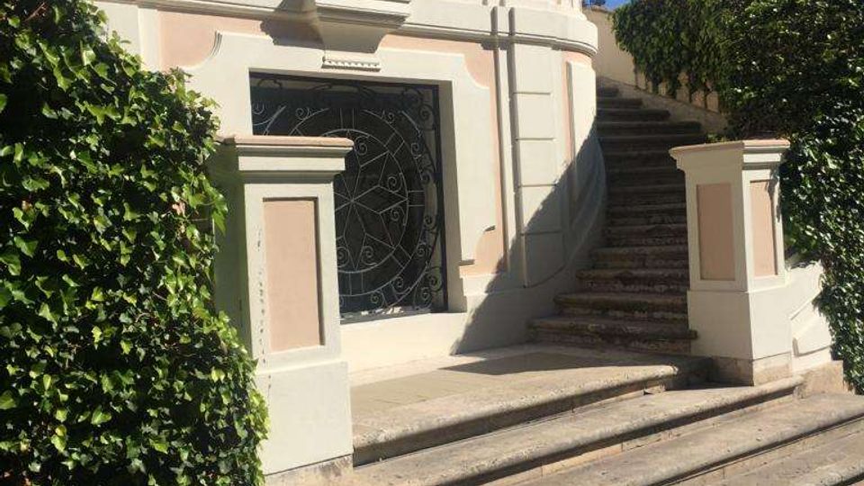 Facciata di una villa in stile classico con pareti rosa e bianche, balaustra e grandi vasi di terracotta. In primo piano una scalinata in pietra conduce a un ingresso con cancellata in ferro battuto, affiancata da siepi e palme in giardino.