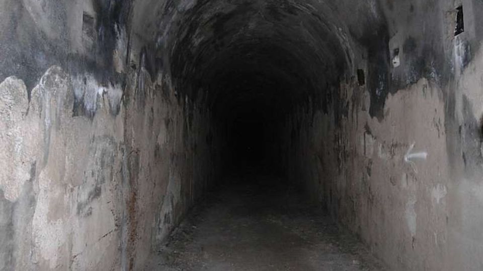 Interno di un lungo tunnel sotterraneo con volta a botte, pareti in cemento macchiate e annerite e pavimento polveroso. Piccole aperture laterali interrompono le pareti mentre il corridoio si perde nel buio in fondo.