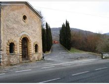 Piccola cappella in pietra con portale e finestre ad arco e oculo sulla facciata, affacciata su una strada asfaltata. Accanto parte un viale in lieve salita fiancheggiato da cipressi che conduce verso edifici e colline boscose sullo sfondo.