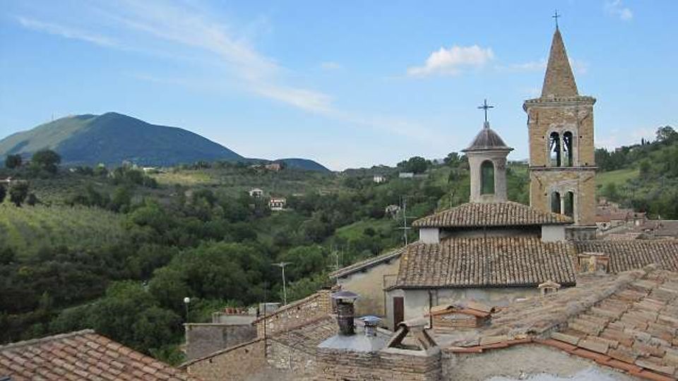 Veduta panoramica di un borgo collinare con tetti in coppi e una chiesa in pietra con campanile e piccola torretta. Sullo sfondo si estendono colline e un monte verde sotto un cielo sereno.