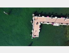 Veduta dall’alto di un molo a forma di L con pavimentazione rosa e ringhiere, decorato con fioriere, che si estende su acqua verde scura. Si notano alcune piccole imbarcazioni e le ombre delle strutture riflesse sull’acqua.