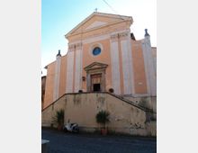 Facciata di una chiesa in stile classico con timpano triangolare e croce in cima, grandi paraste verticali e rosone circolare sopra il portale. Una scalinata con muretto conduce all’ingresso, con vasi di piante e strada lastricata in primo piano.