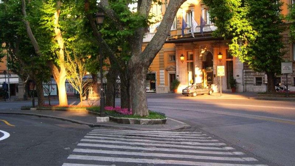 Attraversamento pedonale a strisce bianche su una strada urbana con spartitraffico alberato e aiuole fiorite. Sullo sfondo si vede un edificio storico color ocra con balconi e un grande ingresso ad arco illuminato da lampioni.