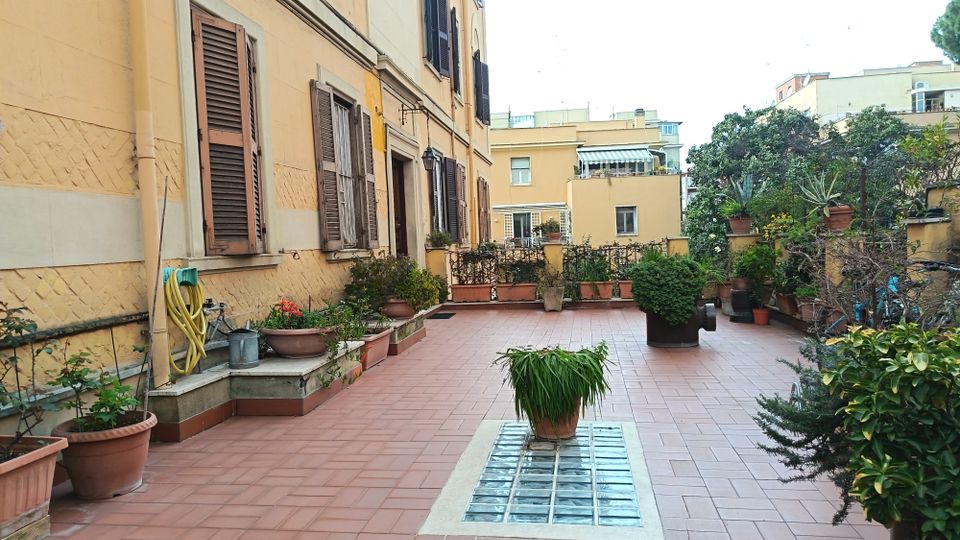 Ampia terrazza condominiale con pavimentazione in mattoni rossi, fioriere e vasi di piante lungo i lati e una griglia di vetro al centro. A sinistra si vede la facciata di un edificio con finestre e persiane, mentre sullo sfondo compaiono altri palazzi e alberi.
