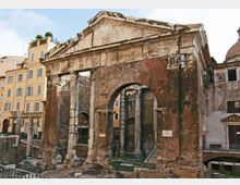 Rovine di una struttura monumentale in mattoni e pietra con grande arco centrale e colonne parzialmente conservate, affacciata su una piazza urbana. Sullo sfondo si vedono edifici residenziali con facciate chiare e una cupola.